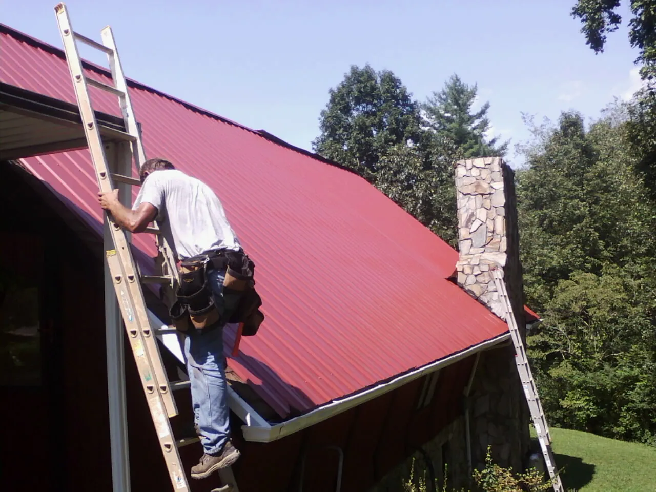 Roofer on a ladder beside a red metal roof and stone chimney — Soffit & Fascia Repair by Roofing Brothers in Atlantic Beach